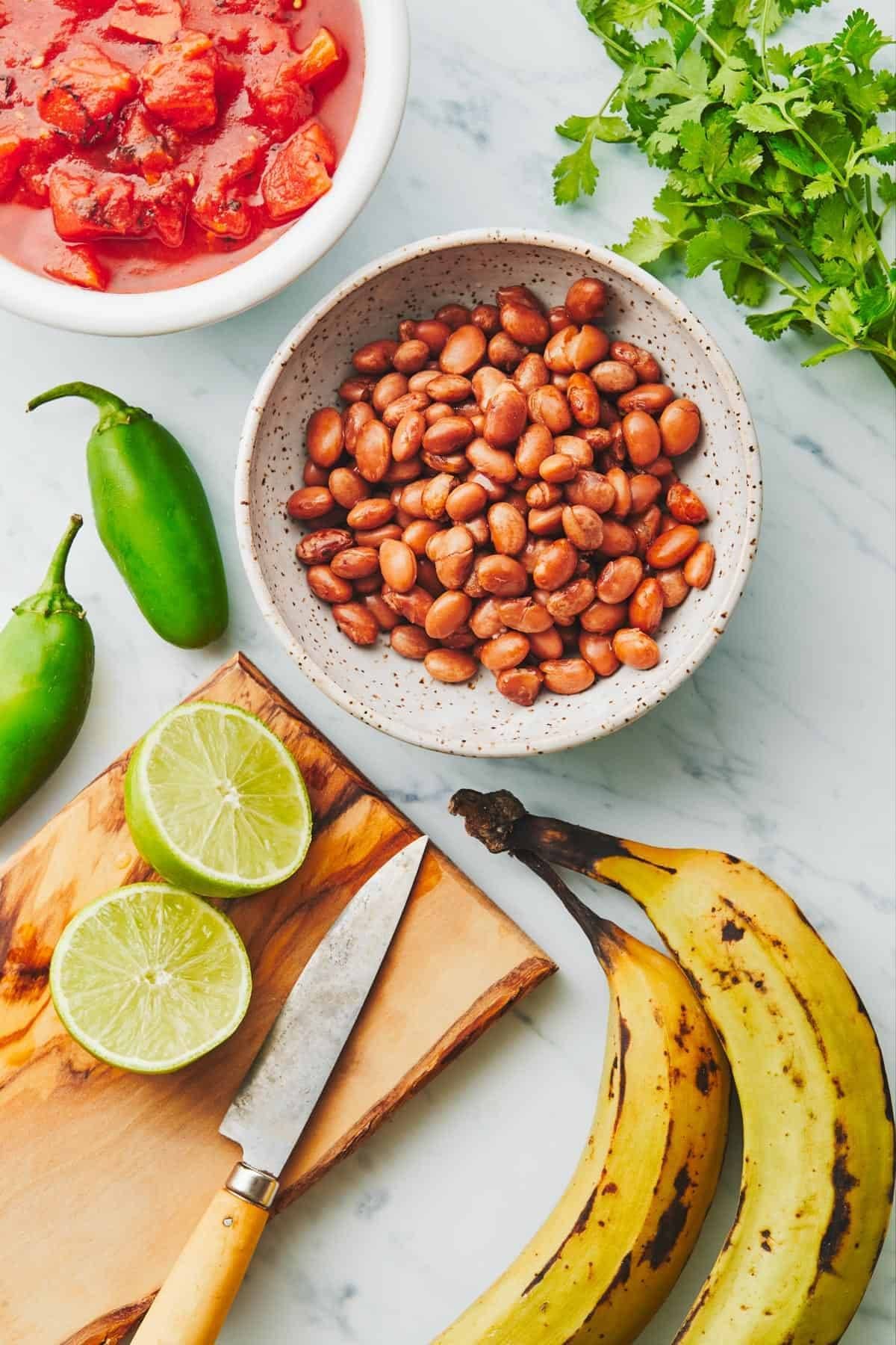 Tomatoes, pinto beans, jalapeños, lime, plantains, and cilantro on a cutting board.