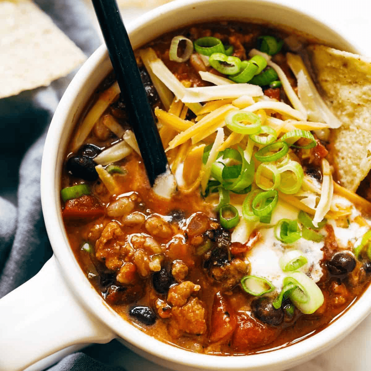 Turkey chili in a bowl with cheese, green onion, sour cream, and tortilla chips.